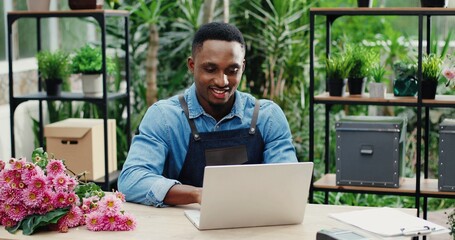 Joyful young man in flower shop sitting at workplace and texting on computer. Cheerful African...