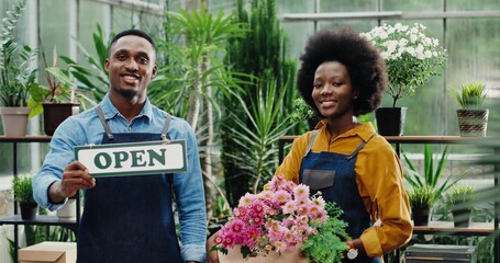 Portrait of African American happy male florist in apron standing at flower shop and smiling to camera with Open sign in hand. Female entrepreneur rholding box with beautiful flowers Reopening concept