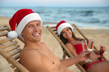 Obraz premium Happy couple wearing Santa hats and drinking champagne together on beach. Christmas vacation