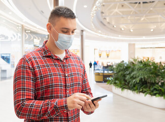 A young man in a medical mask in a shopping center looks into his smartphone. Covid-19