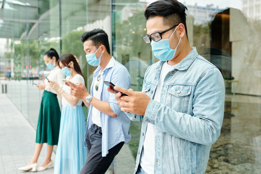 Young Asian people wearing medical masks due to coronavirus pandemic, they are standing outside the shopping mall and texting friends or using social media