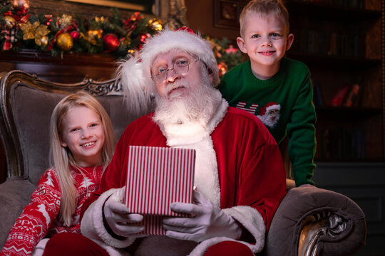 Portrait Santa Claus And Children. Blonde Girl And Boy Opening Christmas Gift Near Christmas Tree.Brother And Sister Celebrating Family Holiday. Magic Fulfillment Of Desires Wishes.