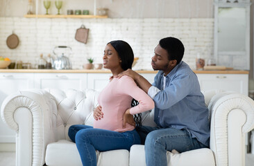 Black expectant woman suffering from back pain, loving husband massaging her shoulders at home