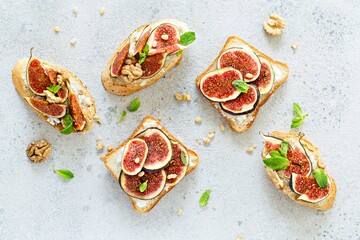Crispy bruschetta with soft ricotta, ripe figs, walnuts and pine nuts, mint and honey on a light background. Figs fruit toast on marble table with honey and walnut. Top View.