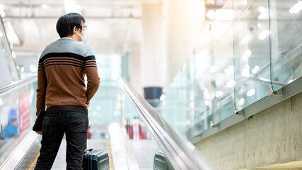 Travel insurance concept. Asian man tourist wearing face mask carrying suitcase luggage and digital tablet on travelator or moving walkway in airport terminal.
