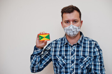 Man in checkered shirt show Grenada flag card in hand, wear protect mask isolated on white background. American countries Coronavirus concept.