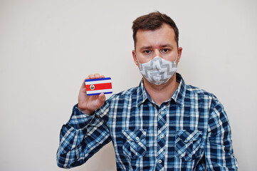 Man in checkered shirt show Costa Rica flag card in hand, wear protect mask isolated on white background. American countries Coronavirus concept.