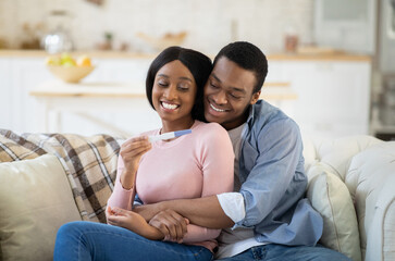 Beautiful black couple holding positive pregnancy test and hugging on sofa at home