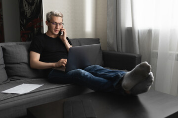 A man with glasses on the couch at home is working on a laptop and talking on the phone. Remote work concept during a pandemic