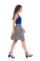 Serious Young Woman In Striped Skirt And High Heels Is Walking. Side View.
