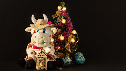 White soft toy bull on a dark background with a Christmas tree and garlands, New Year's card. Chinese Year of the Ox, zodiac symbol 2021