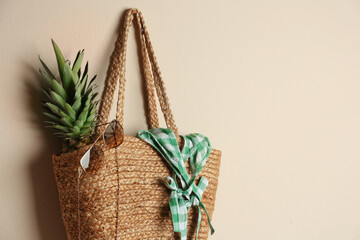 Elegant woman's straw bag with pineapple and accessories hanging on beige background. Space for text