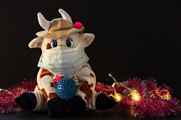White soft toy bull in a medical mask on a dark background with a Christmas tree and garlands, New Year's card. New Year and Christmas during the coronavirus pandemic