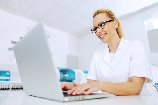 Attractive smiling positive blond lab assistant sitting in laboratory and processing data. - Powered by Adobe