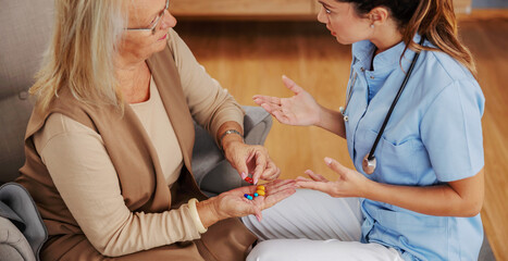 Blond senior woman sitting at home and holding hand full of pills. Nurse sitting next to her and...