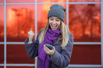 Happy casual excited woman celebrating success holding mobile phone standing on the street warmly dressed in a gray hat and a purple scarf near a shopping center on a bright red background