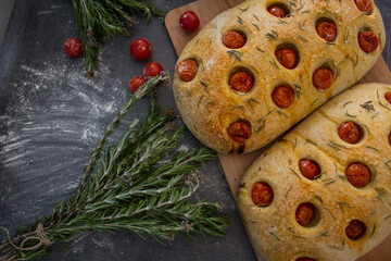 Fresh baked homemade focaccia bread with rosemary and cherry tomatoes on grey textured background. Top view photo. Traditional cooking concept. 
