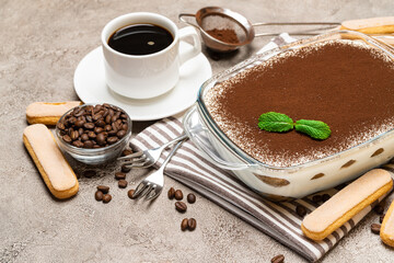 Traditional Italian Tiramisu dessert in glass baking dish, savoiardi cookies and cup of fresh hot espresso coffee on concrete background