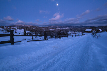 Night countryside road, hoases, hills, groves and farmlands in winter remote alpine mountain village. Ukraine, Voronenko, Moon above village.