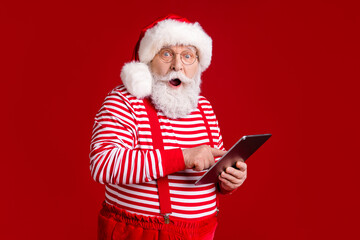Portrait of his he handsome cheerful amazed stunned bearded Santa father holding in hands using device e-book gadget fast speed 5g blog isolated bright vivid shine vibrant red color background - obrazy, fototapety, plakaty