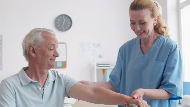 Tilt up of senior Caucasian man sitting on examination couch with dumbbell in his hand while female doctor helping him rasing his arm