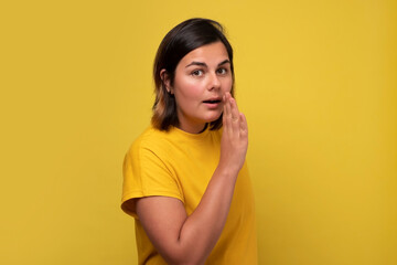 Caucasian woman covering mouth with hand tells secrets to someone, isolated on yellow wall.