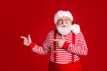 Fototapeta na wymiar Photo of retired old man white beard direct finger empty space suggest interesting solution wear santa x-mas costume suspenders spectacles striped shirt cap isolated red color background