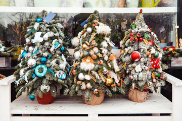Small decorated Christmas trees decorated are on sale in flower shop window. Wreaths and decoration for New Year celebration. Winter holiday spirit.