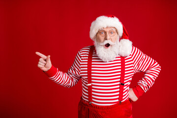 Fototapeta na wymiar Photo of retired old man grey beard direct finger empty space open mouth shock suggest product wear santa costume suspenders spectacles striped shirt headwear isolated red color background