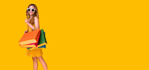 Picture of a shocked young brunette woman in yellow summer dress wearing sunglasses posing with shopping bags and looking at camera over orange background.