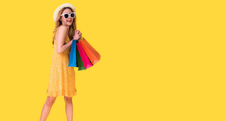 Picture of a shocked young brunette woman in yellow summer dress wearing sunglasses posing with shopping bags and orange background.