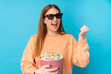 Young redhead woman over isolated background with 3d glasses and holding a big bucket of popcorns