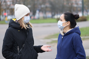 Two women in medical protective masks communicate in street. Safe walks in coronavirus pandemic concept