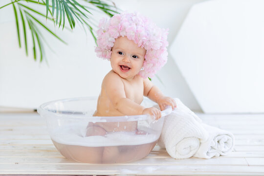 happy little baby girl washes in a basin with foam and water in a bright room at home in a beautiful bathing cap made of flowers