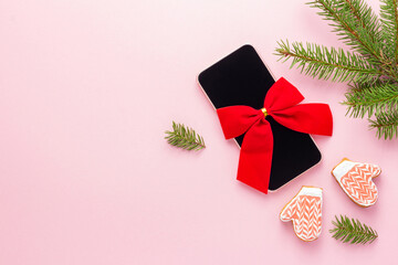 Smartphone with red bow like a Christmas present on pink background next to spruce and cookies