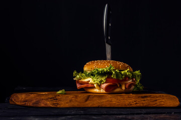 Homemade burger with ham, cheese, salad and pomegranate, pierced by a knife on a wooden board