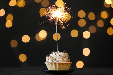 Cupcake with burning sparkler on black table against blurred festive lights