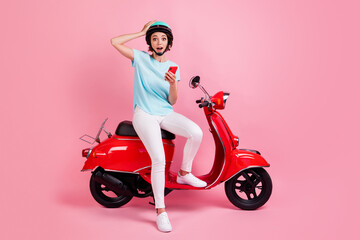 Fototapeta na wymiar Photo of astonished pretty young woman sit moped hold telephone open mouth wear pants shoes slam t-shirt isolated on pink background