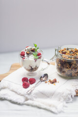 Granola in a jar and in a cup with yogurt and raspberry on white background. Granola is a breakfast food and snack food consisting of rolled oats, nuts, honey, that is baked until golden brown
