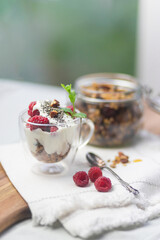 Granola in a jar and in a cup with yogurt and raspberry near window, green background. Granola is a breakfast food and snack food consisting of rolled oats, nuts, honey, that is baked until brown