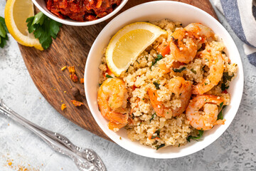 Couscous with fried shrimps, tomatoes, onions, and herbs in a white plate on a gray concrete table top view. Selective focus. Tasty Moroccan food with couscous and seafood.