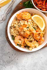 Couscous with fried shrimps, tomatoes, onions, and herbs in a white plate on a gray concrete table top view. Selective focus. Tasty Moroccan food with couscous and seafood.