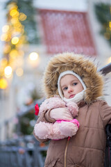 A girl in a white hat, a hood with fur and a brown jacket stands on the street and hugs a bear. Christmas holiday street with lights