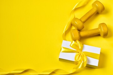 Sport flat lay composition with yellow dumbbells and gift box with yellow bow on yellow background. Top view. copy space. Christmas sport equipment. Concept holiday.