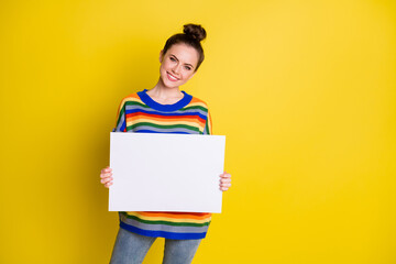 Photo of cheerful young lady wear rainbow striped sweater hold white card recommend ad isolated on yellow color background