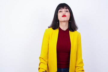 Attractive Caucasian woman standing against white background wearing yellow blazer rounds lips,...