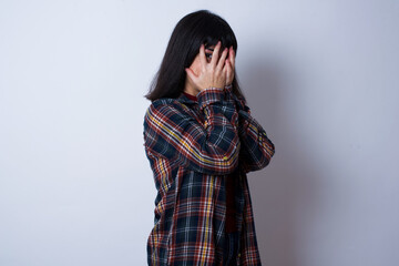 Beautiful young Caucasian woman standing against white background covering face with hands and peering out with one eye between fingers. Scared from something or someone.