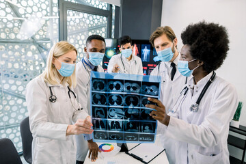 Multiethnical five doctors, wearing lab coats and medical masks, examining an x-ray CT scan image at modern hospital to make diagnosis and treatment. Radiology, pulmonology oncology concept