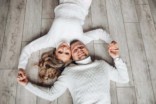 Happy loving couple in white knitted winter sweaters lying on the floor holding hands. From above.
