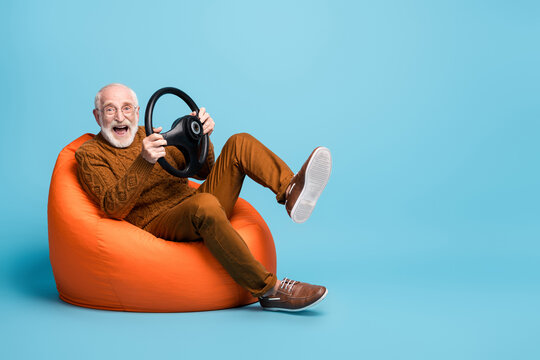 Portrait of his he nice attractive cheerful cheery glad funny bearded grey-haired man sitting in bag chair having fun holding in hands steering wheel isolated over blue pastel color background